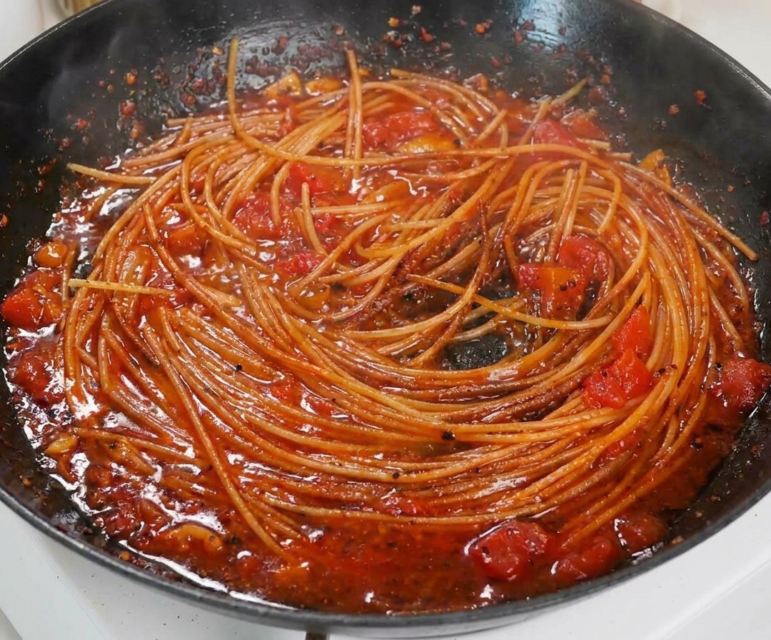 Primer plano de espaguetis cocinados mezclados en una rica y espesa salsa de tomate en una sartén negra.