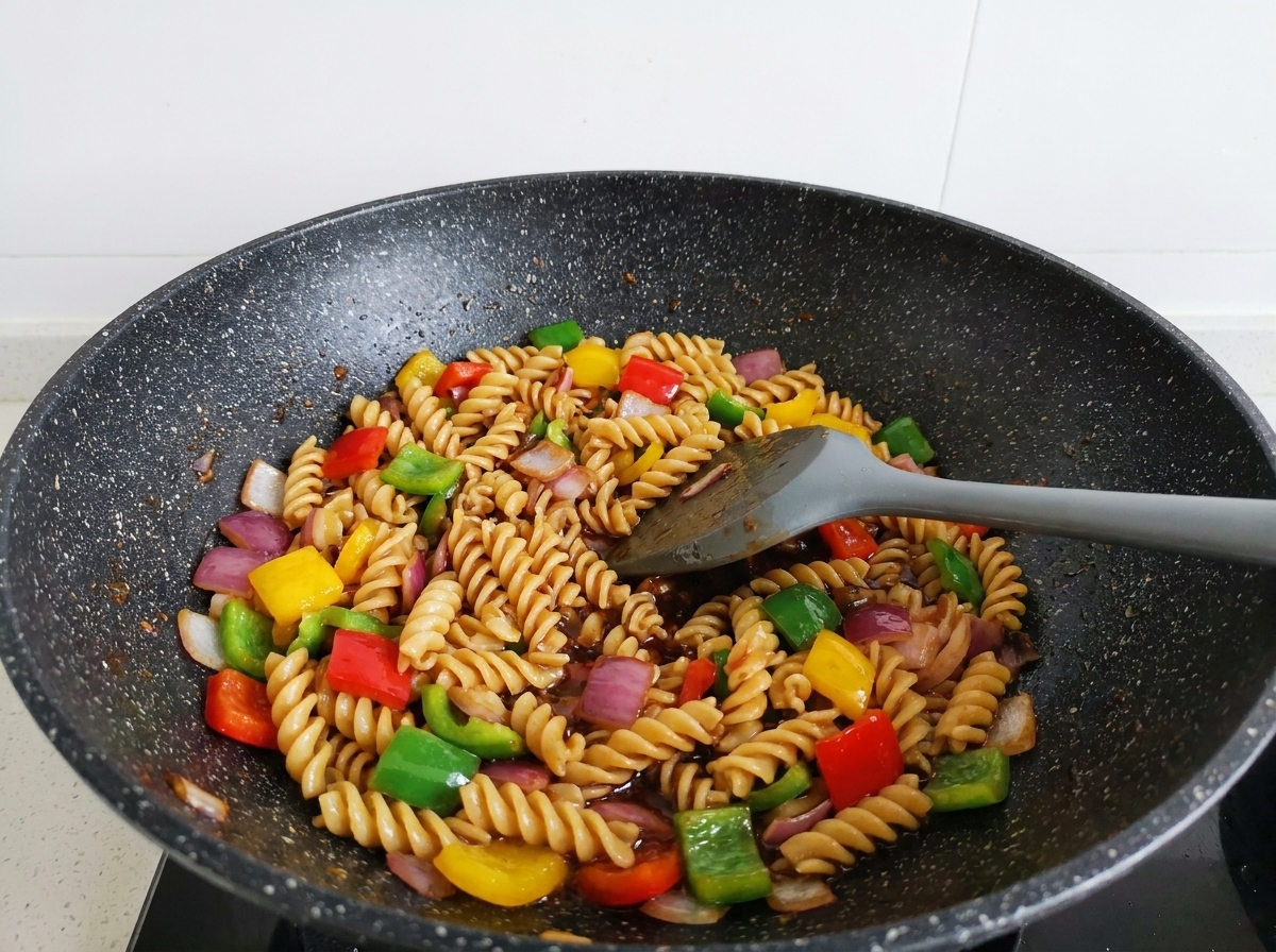 Pasta rotini, tiras de ternera y pimientos de colores salteándose con una espátula gris en un wok, completamente cubiertos por una brillante salsa oscura.