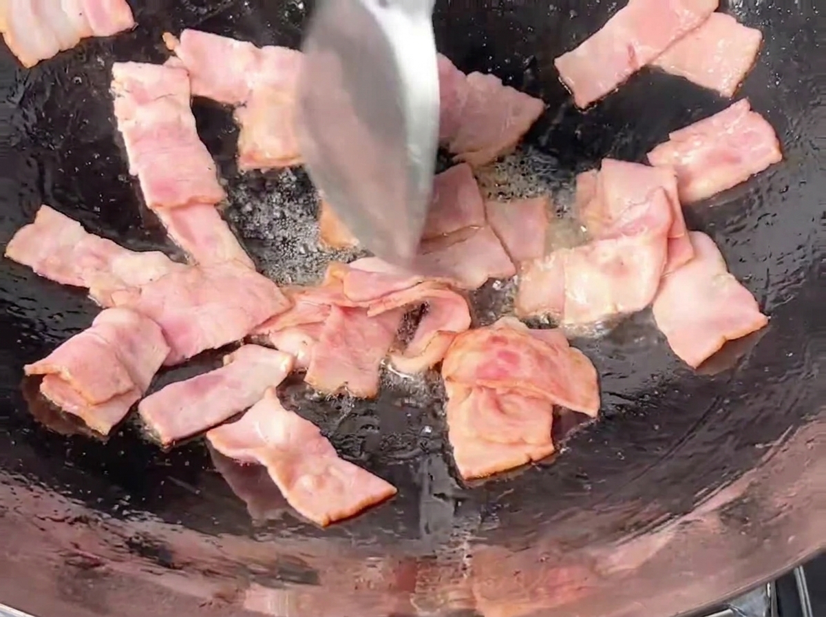 Trozos rectangulares de beicon rosa siendo salteados en un wok de hierro oscuro con una espátula de metal.