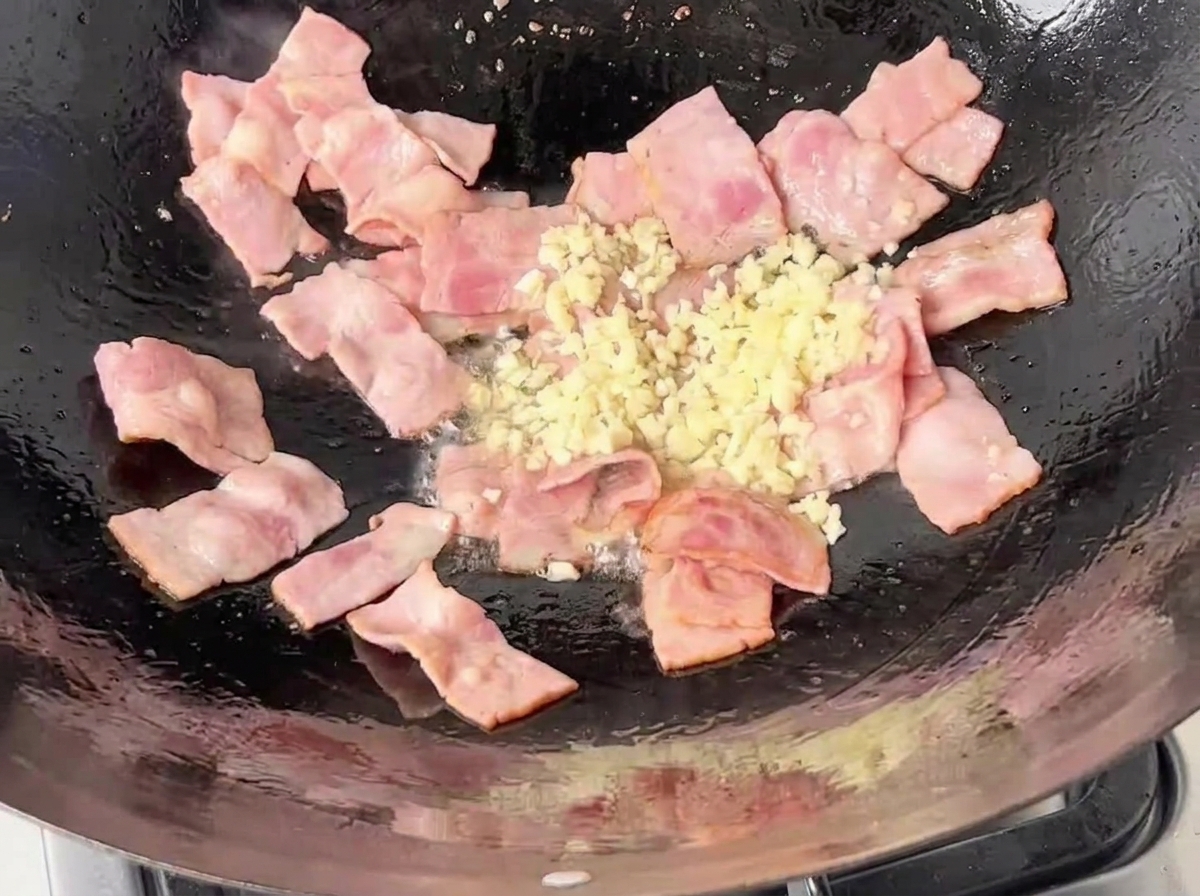 Ajo blanco picado finamente siendo añadido a la sartén con trozos de beicon a medio cocinar.