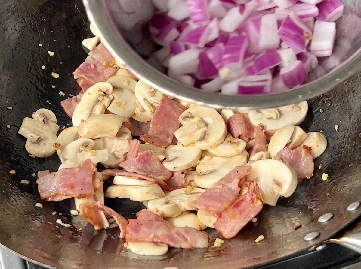 Un wok lleno de champiñones, trozos de beicon y cebolla morada picada siendo salteados juntos.