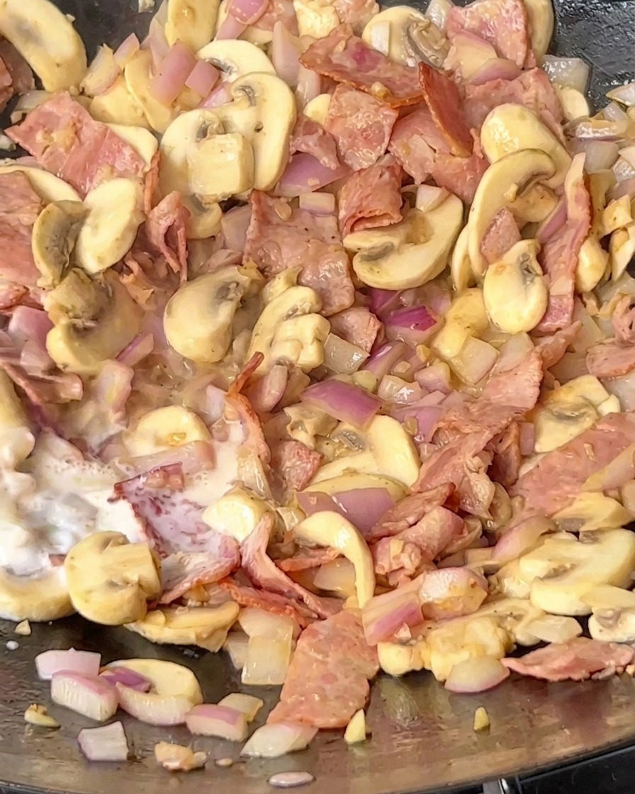 Leche siendo vertida en una sartén que contiene beicon, champiñones y cebolla.