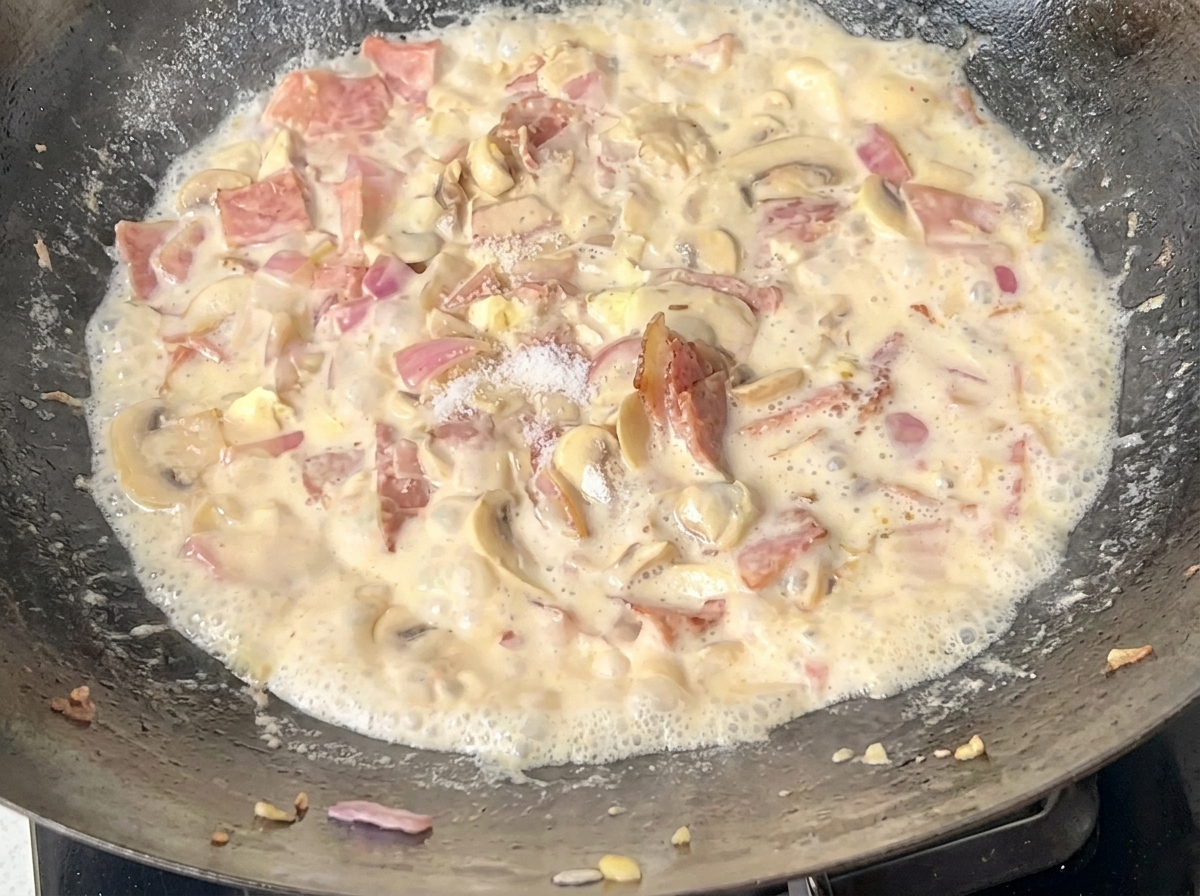 Espaguetis cocidos siendo añadidos a una sartén llena de salsa cremosa de beicon y champiñones.