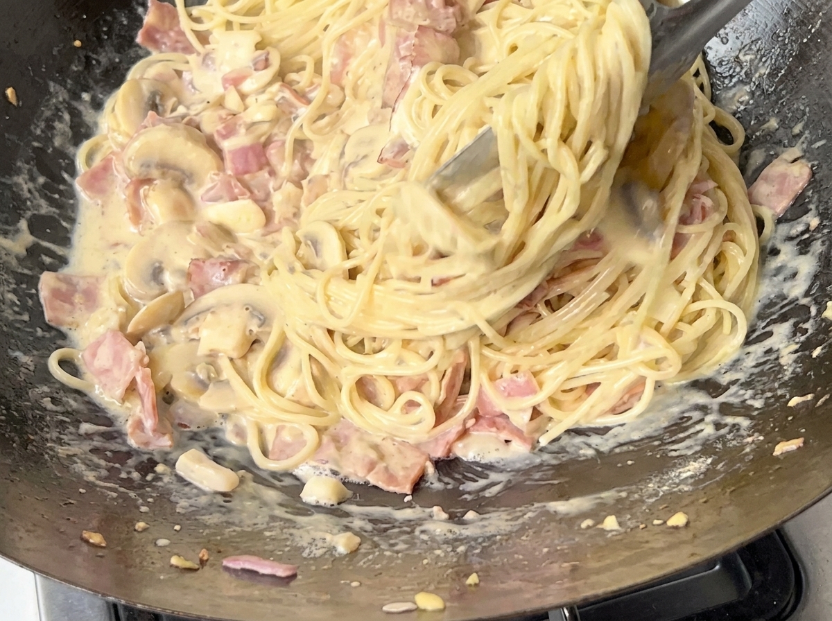 Un primer plano de espaguetis siendo mezclados en una salsa cremosa de beicon y champiñones con pinzas de metal.