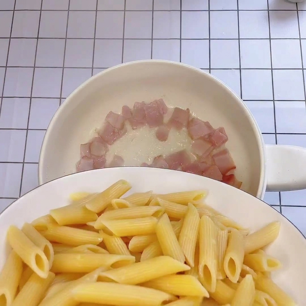 Penne recién cocido siendo añadido a la sartén con trozos de tocino crujiente.