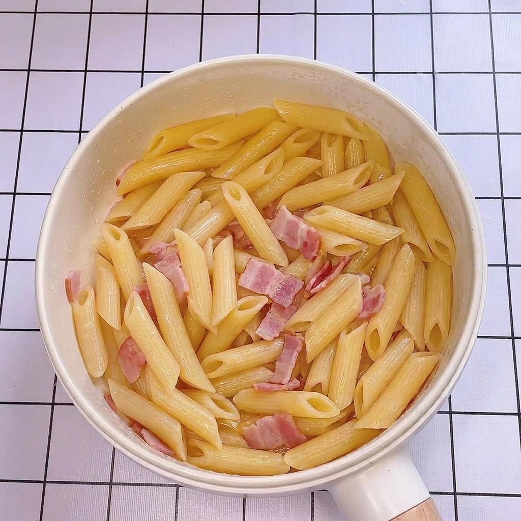 Agua de la pasta siendo vertida en la sartén con penne y tocino, creando un brillo satinado.
