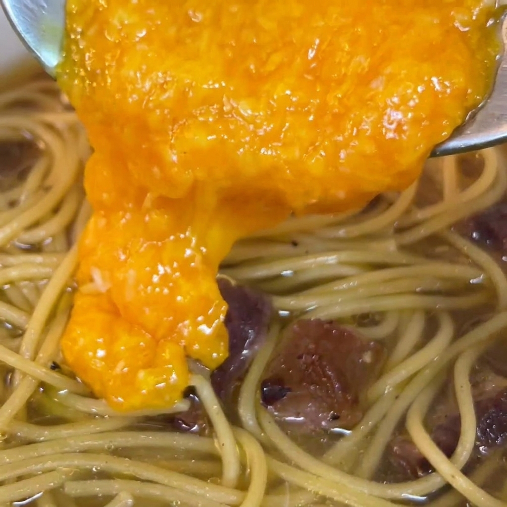 Una mezcla rica de huevo y queso de color naranja vibrante siendo vertida desde una cuchara sobre la pasta en la sartén.