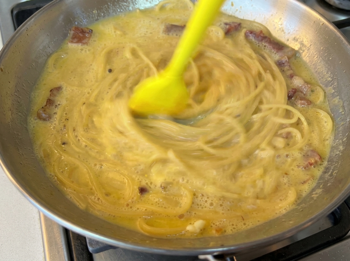 Una espátula de goma amarilla mezclando rápidamente los espaguetis con una salsa cremosa a base de huevo en una sartén de acero inoxidable.