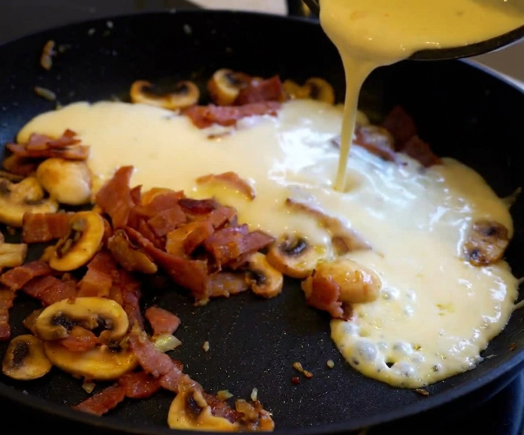 Pasta cocida siendo añadida a una sartén con salsa cremosa de beicon y champiñones.