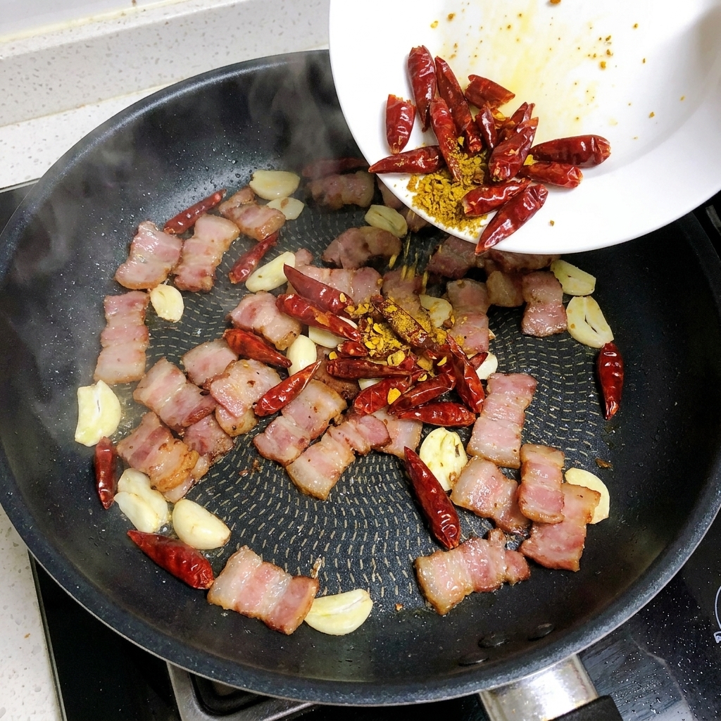 Láminas de ajo y chiles rojos siendo añadidos a una sartén con tocino.