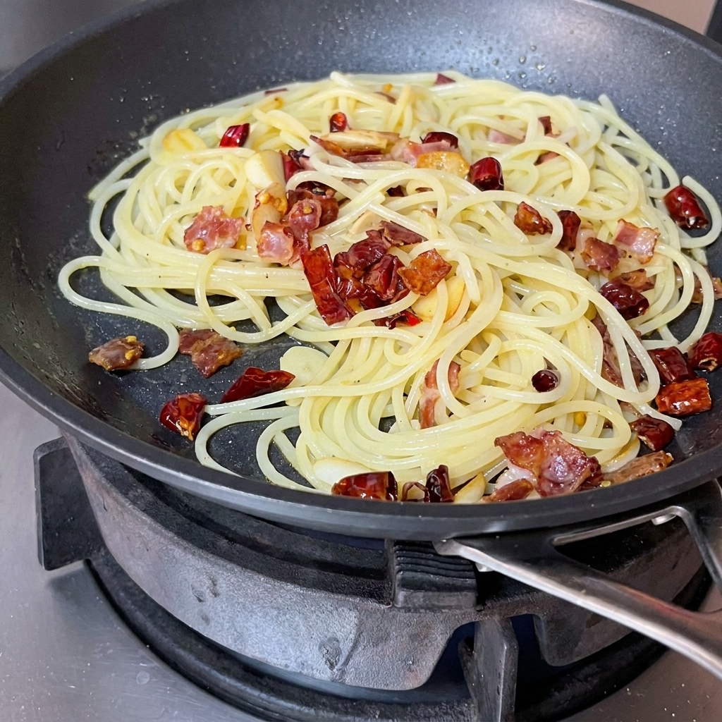 Espaguetis, tocino y aromáticos siendo mezclados en una sartén con una espátula roja.
