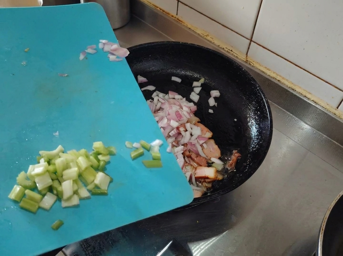 Chalotas picadas y apio siendo añadidos a la sartén con el bacon dorado.