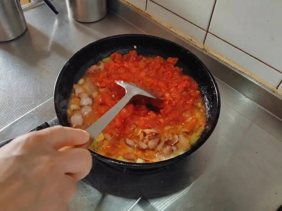 Una espátula de metal aplastando tomates troceados en una sartén mientras se cocinan con bacon y aromáticos.