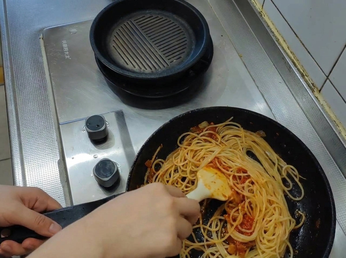 Un cucharón vertiendo agua turbia y hirviendo de la pasta en una sartén llena de espaguetis y salsa de tomate.