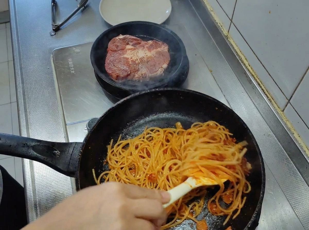 Espaguetis siendo mezclados con pinzas en una sartén, quedando completamente cubiertos por una vibrante salsa de tomate.