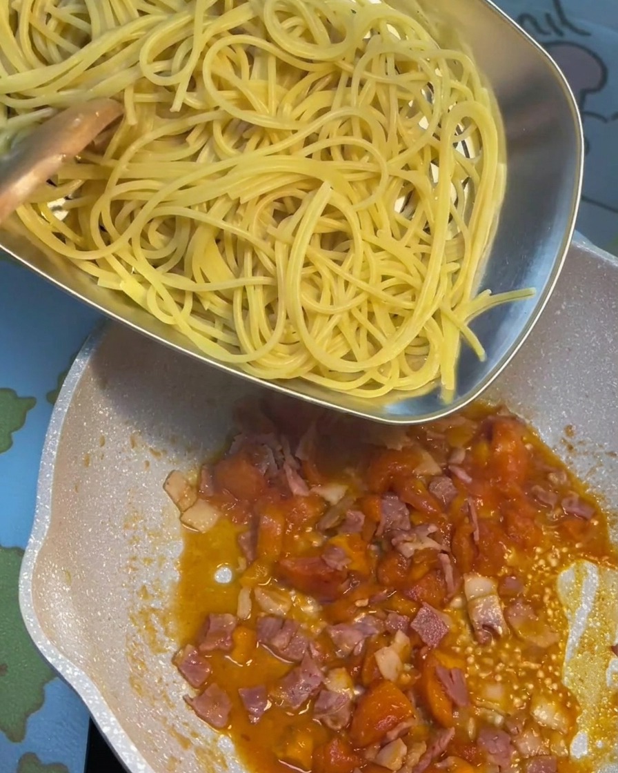 Pasta cocida siendo mezclada en una sartén con una rica salsa de tomate y tocino.