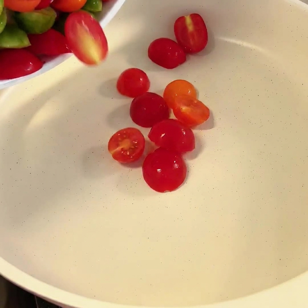 Tomates cherry cortados a la mitad cayendo en una sartén blanca.