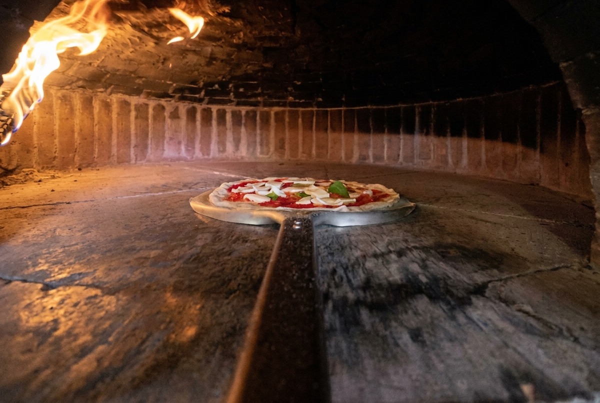 Una pala de metal alargada introduciendo la pizza cruda en un horno de leña muy caliente.