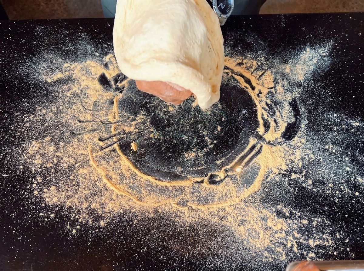 Mano estirando una porción de masa de pizza en el aire sobre una superficie negra con harina.