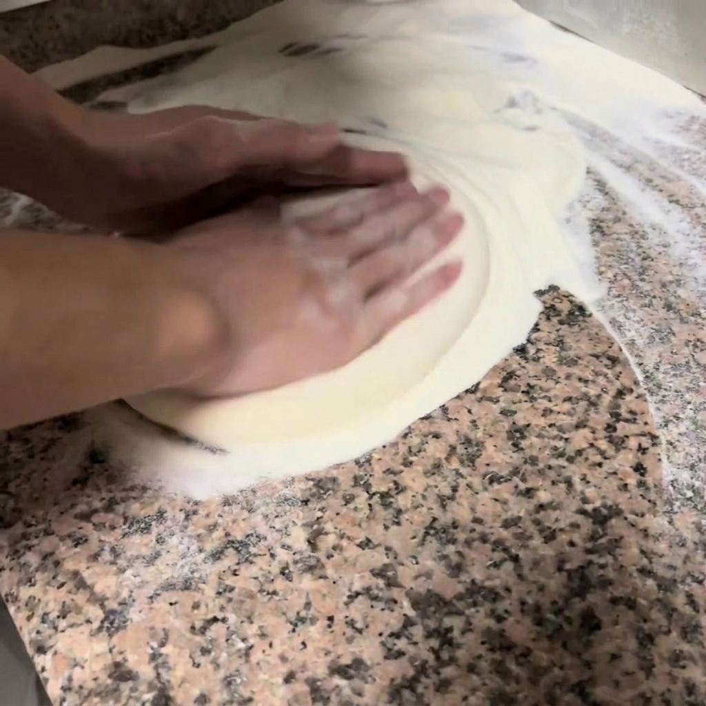 Manos presionando y estirando suavemente una masa de pizza redonda sobre una encimera enharinada.