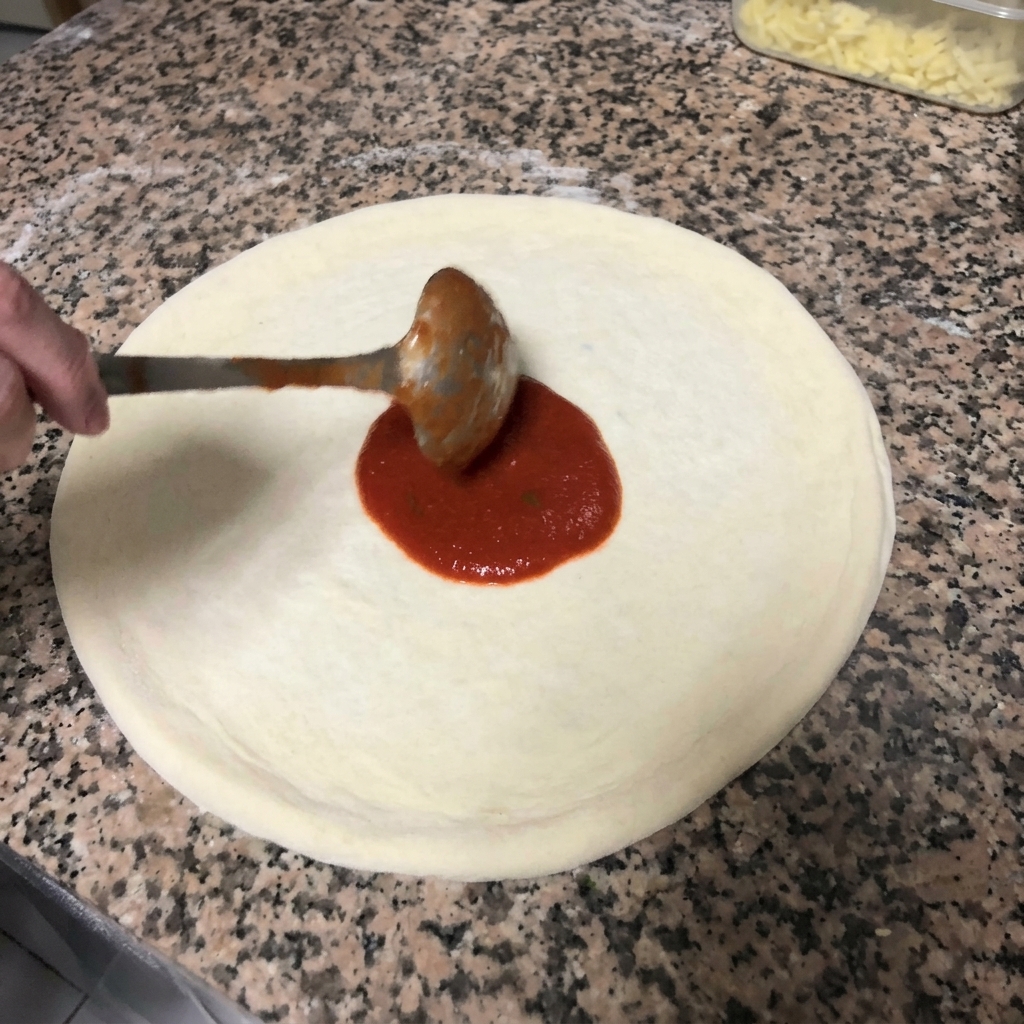 Extendiendo salsa de tomate roja sobre la masa de pizza con movimientos circulares usando un cucharón de metal.