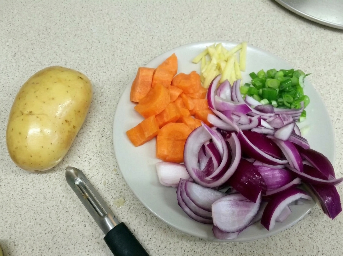 Un plato blanco con zanahorias picadas, cebollas rojas, tiras de jengibre y cebolletas junto a una patata cruda y un pelador.