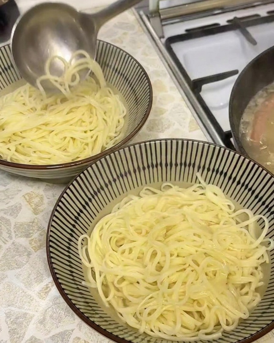 Un cucharón vertiendo caldo de cerdo dorado y opaco en un tazón que contiene fideos de ramen recién cocidos.