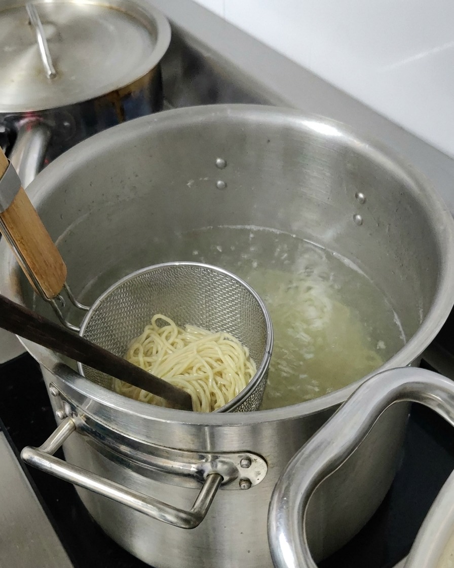 Fideos ramen cociéndose dentro de un colador metálico sumergido en una olla grande con agua caliente.