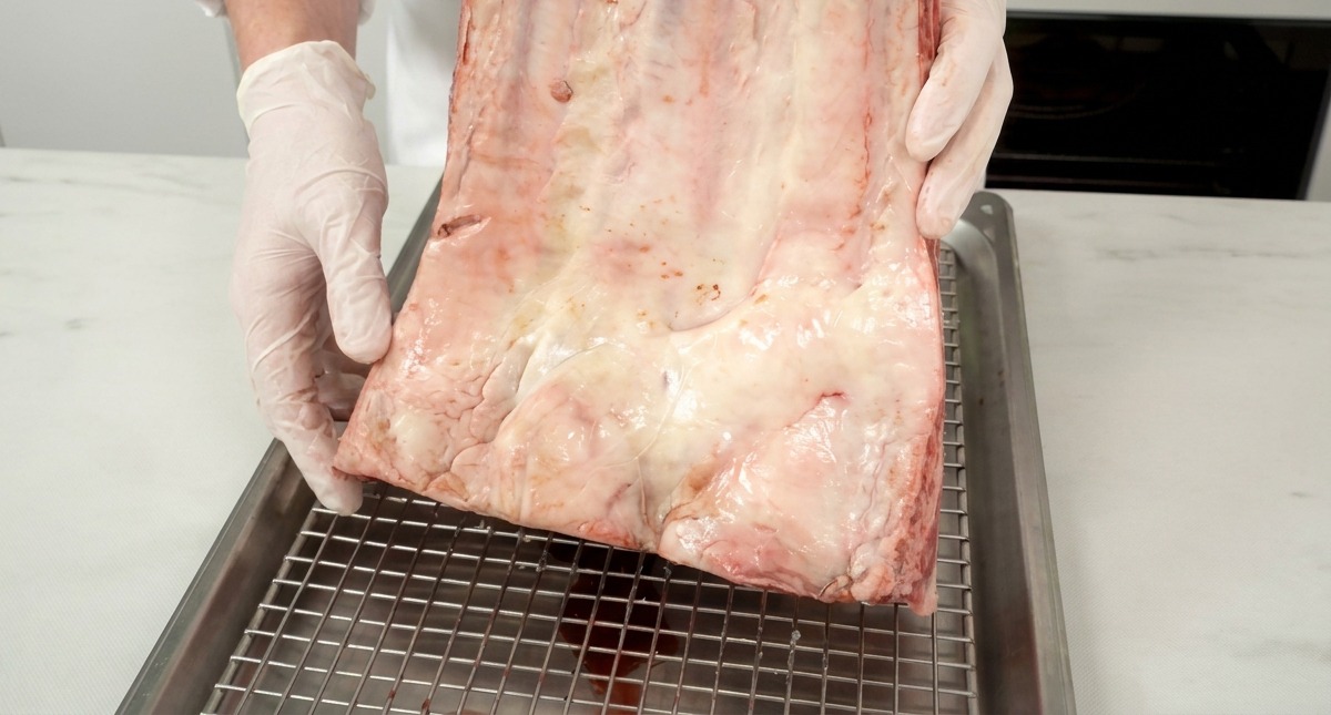 Un trozo grande de costillas de res crudas con marmoleo blanco visible sostenido por un chef sobre una rejilla metálica.