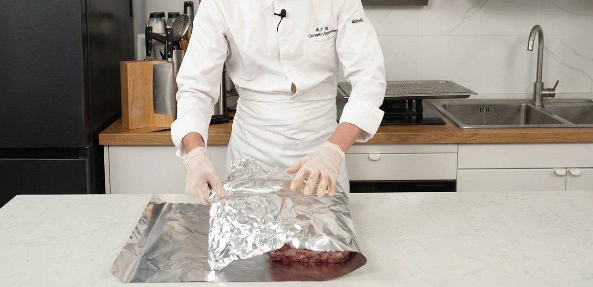 Un chef doblando una hoja grande de papel de aluminio plateado sobre costillas de res sazonadas en una encimera.