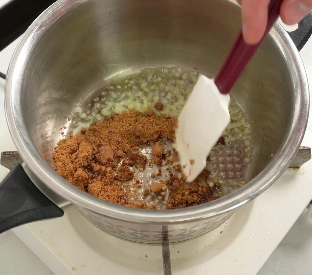 Azúcar morena siendo mezclada en una olla de acero inoxidable con cebollas y ajo sofritos usando una espátula blanca.