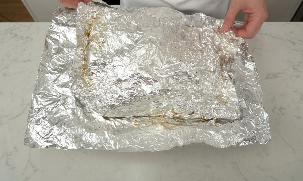 Un par de manos retirando con cuidado el papel de aluminio arrugado para revelar una pieza grande de costillas de res asadas lentamente.