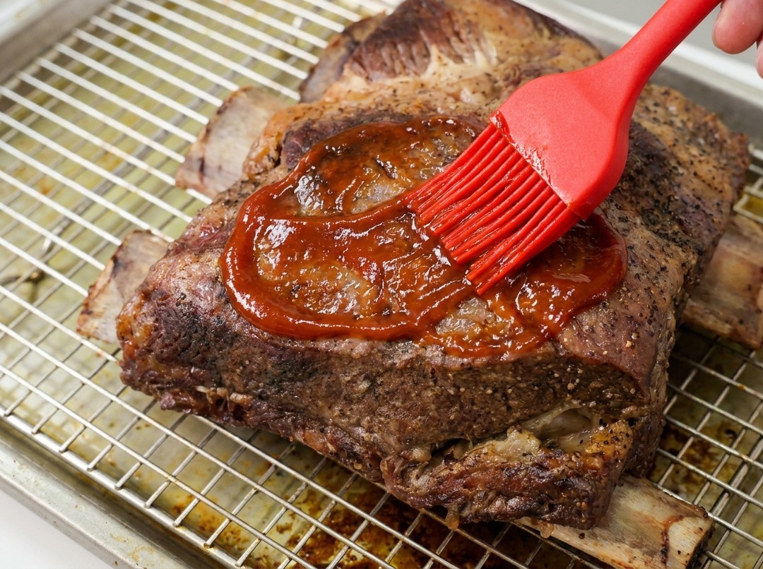 Una mano usando un pincel de silicona rojo para untar una salsa BBQ espesa y brillante sobre costillas de res cocinadas.