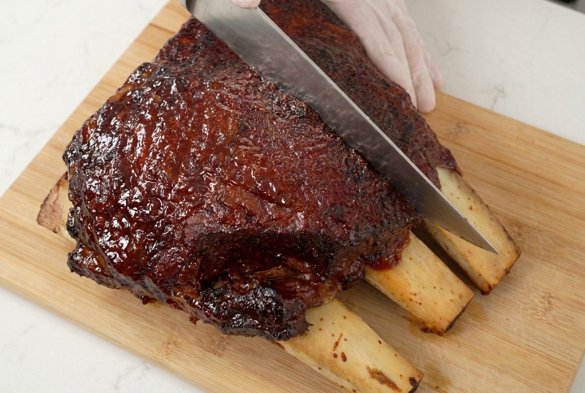 Un cuchillo de chef afilado cortando una pieza de costillas caramelizada sobre una tabla de madera, mostrando la textura jugosa.
