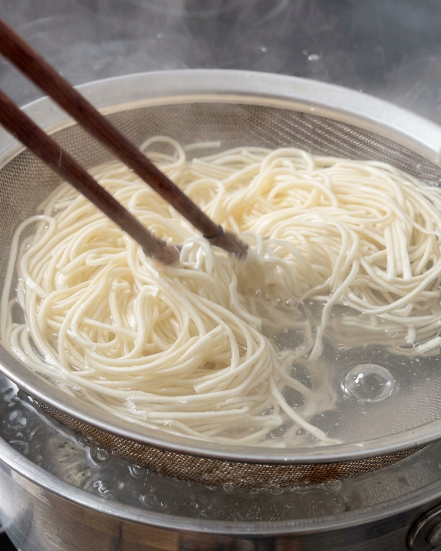 Fideos ramen frescos siendo retirados de agua hirviendo en un colador de metal con palillos de madera.