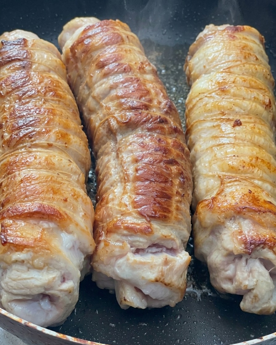Tres rollos grandes de panceta de cerdo atados sellándose en una sartén caliente, mostrando una costra dorada y caramelizada.