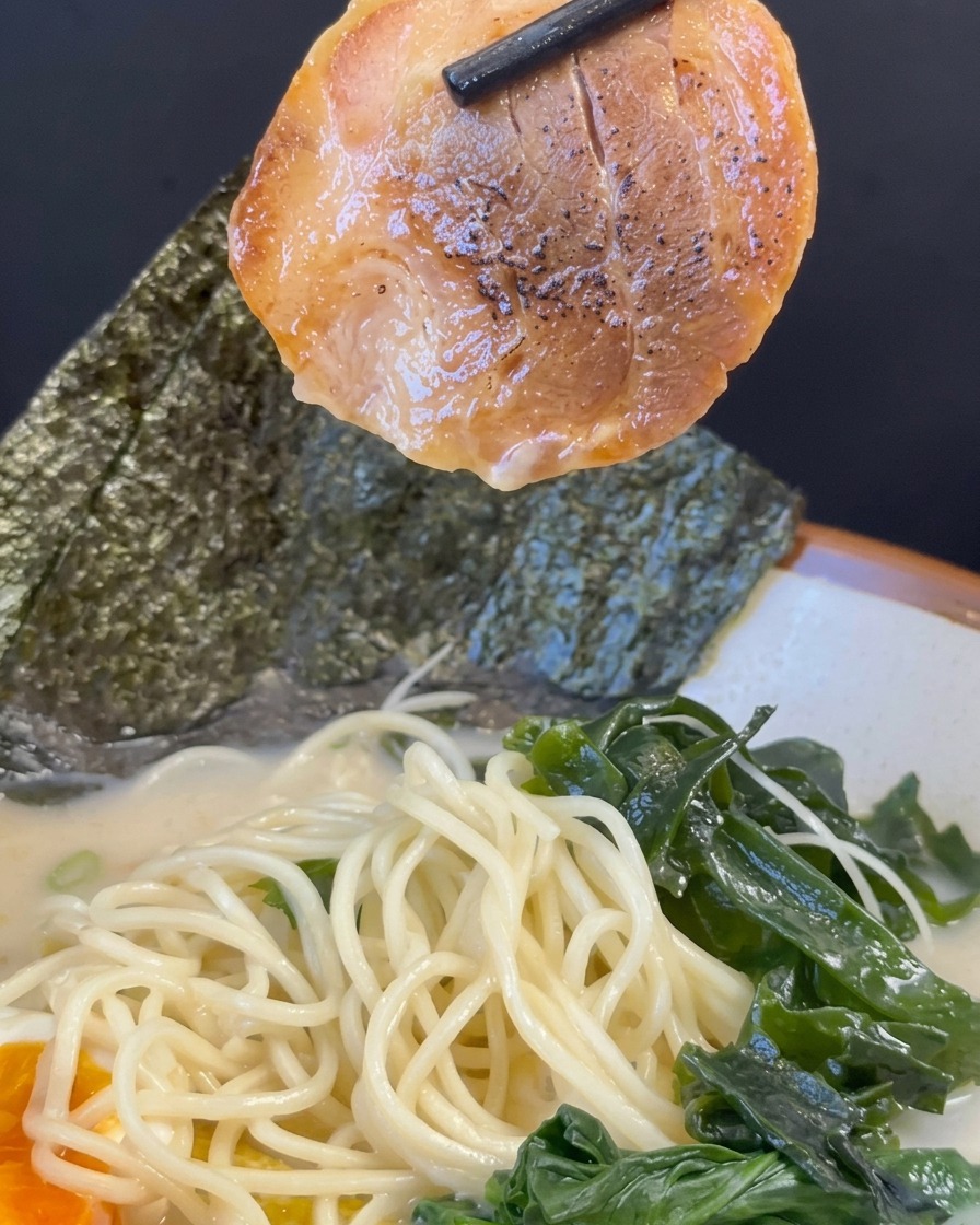 Palillos negros levantando una rebanada de cerdo char siu caramelizado sobre un tazón de ramen ya montado con fideos, caldo, algas y huevo.