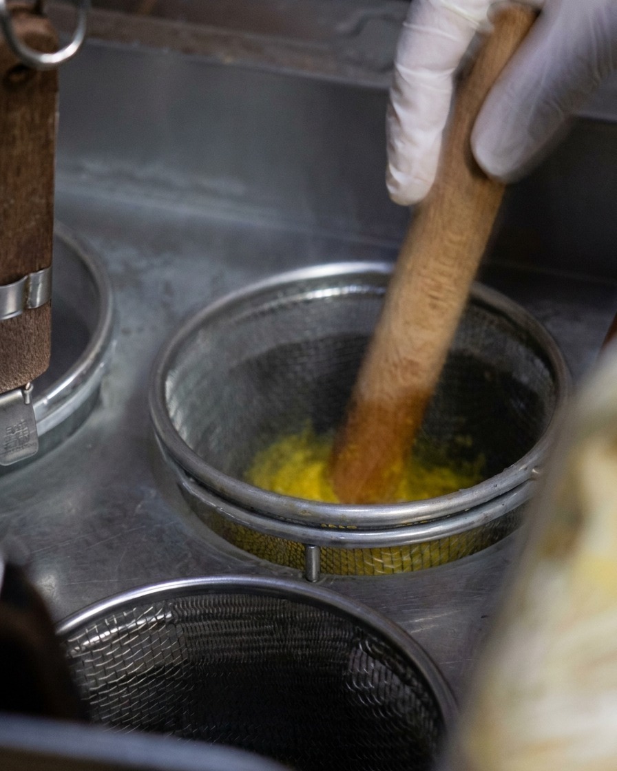 Una cesta de malla metálica con fideos amarillos siendo removidos con una herramienta de madera en una olla de agua hirviendo.