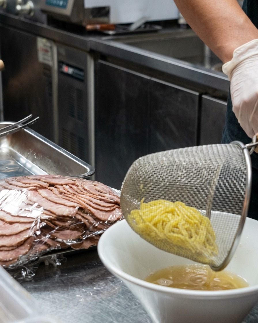 Un colador metálico dejando caer fideos de ramen frescos en un tazón blanco con caldo cremoso.