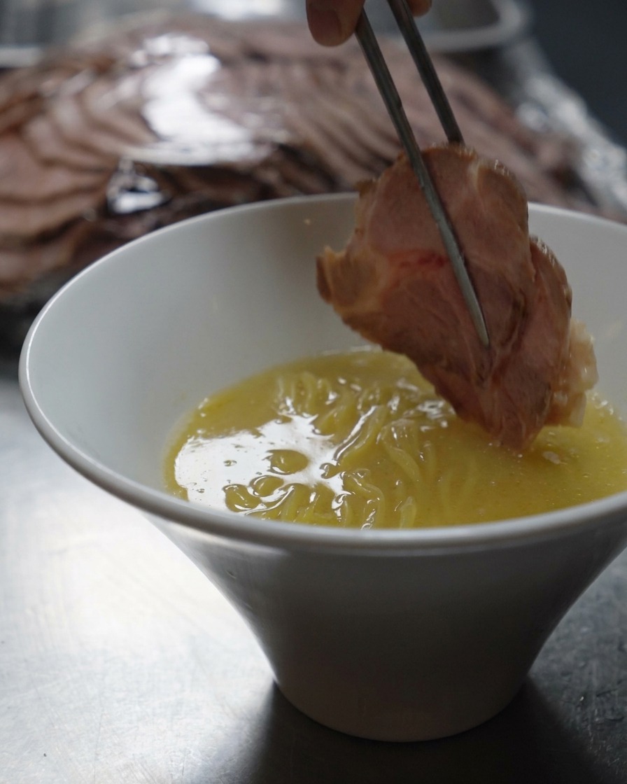 Palillos metálicos colocando una rodaja gruesa de cerdo sellado en un tazón de ramen.