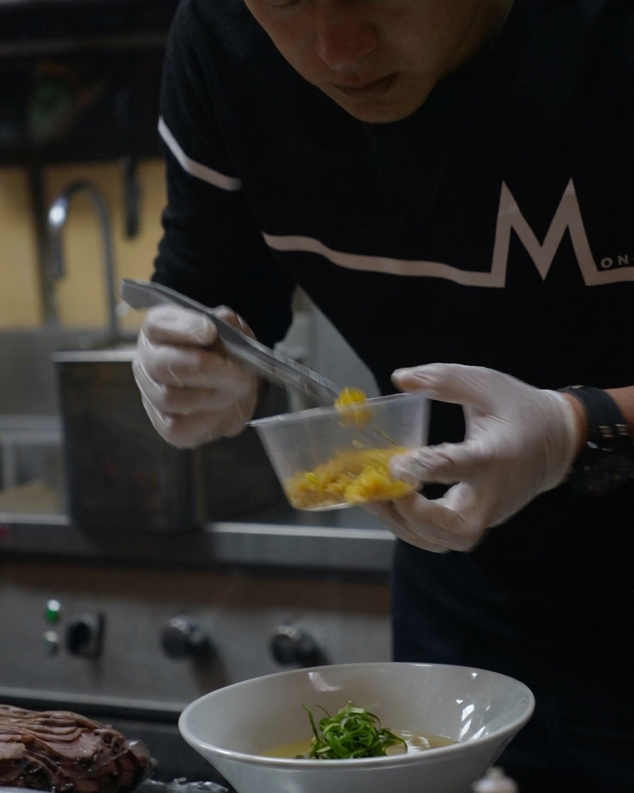 Una pequeña cuchara con ralladura de yuzu amarilla brillante siendo espolvoreada sobre el ramen terminado.