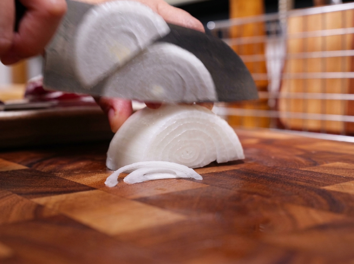 Un cuchillo de chef cortando media cebolla blanca en tiras finas sobre una tabla de cortar de madera.