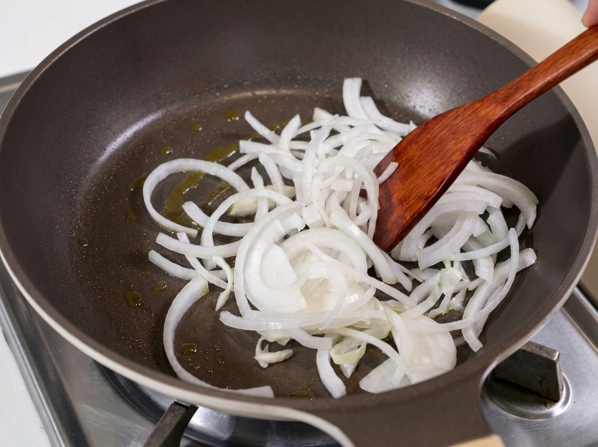 Tiras de cebolla blanca siendo salteadas en una sartén con aceite usando una espátula de madera.