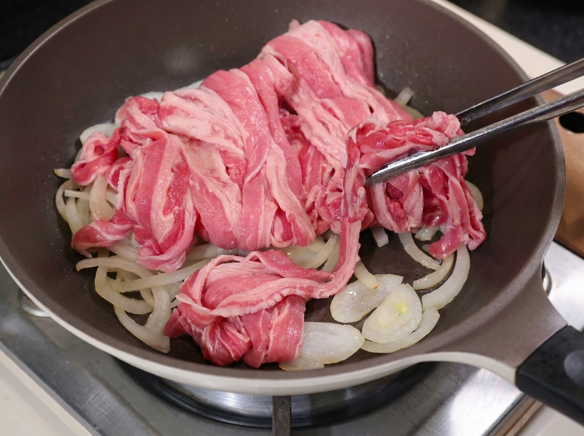 Pinzas metálicas colocando láminas de ternera sobre la cebolla cocinada en una sartén caliente.