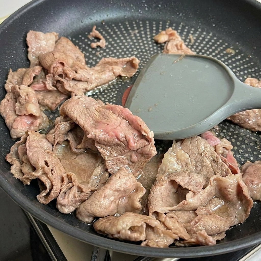 Una espátula levanta tiras de carne tierna y parcialmente cocida de la sartén.