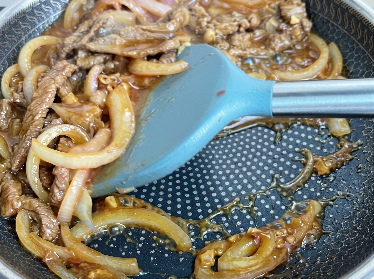 Carne y cebollas removidas en una salsa oscura y burbujeante dentro de una sartén.