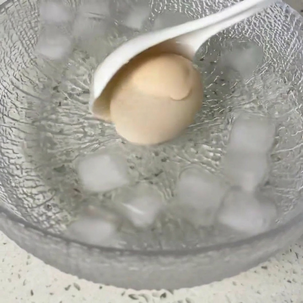 Un huevo cocido siendo trasladado a un tazón de vidrio lleno de cubitos de hielo y agua.