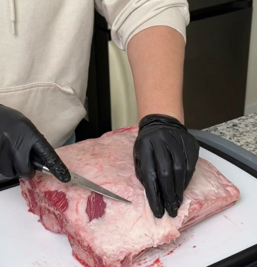 Una persona con guantes negros usa un cuchillo deshuesador para quitar la grasa y la membrana de un costillar de ternera crudo.