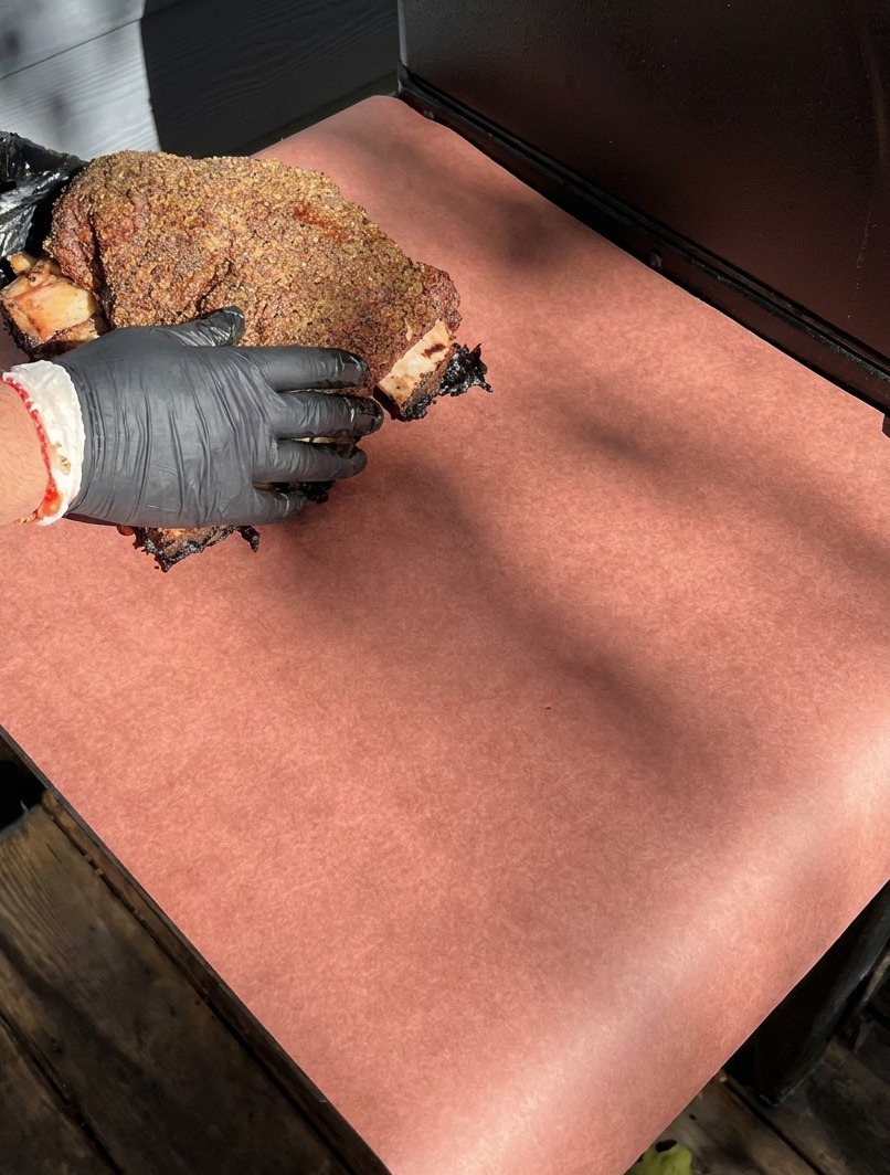 Manos con guantes colocando las costillas de ternera ahumadas sobre una hoja grande de papel de carnicero rosa para envolverlas.