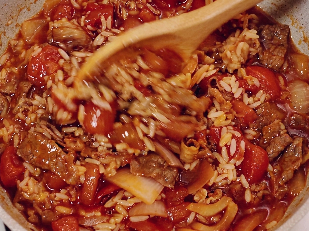 Una cuchara de madera removiendo la ternera, el arroz y la salsa de tomate en una sartén.